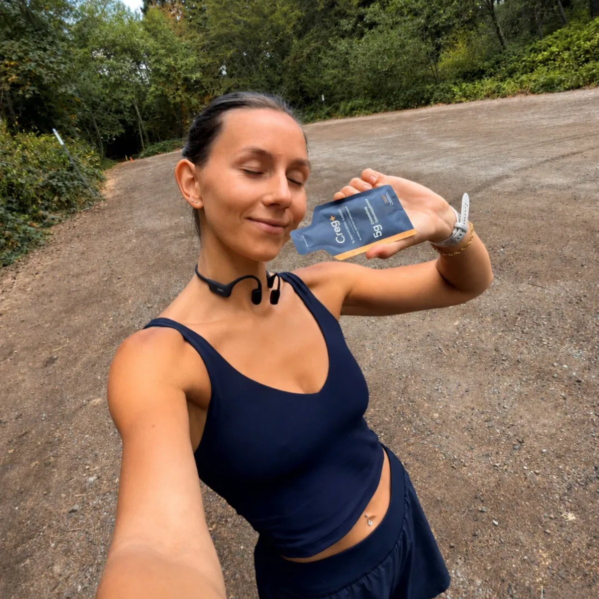 Woman in navy athletic wear holding a CREG+ creatine gel sachet on a tree-lined dirt trail.